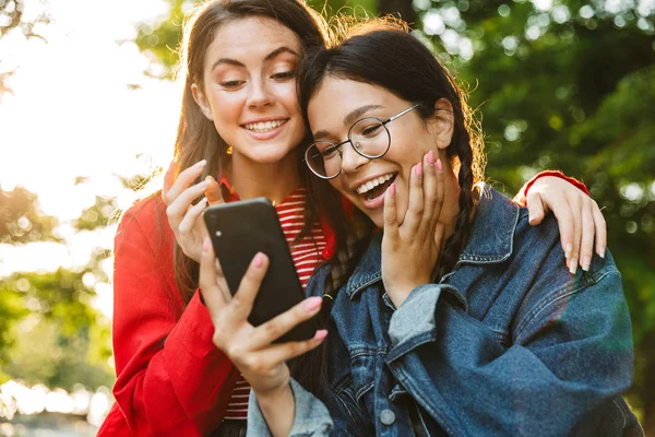 Immagine di due studentesse eccitate che usano il cellulare e si abbracciano mentre siedono sulla ringhiera nel parco verde — Foto Stock