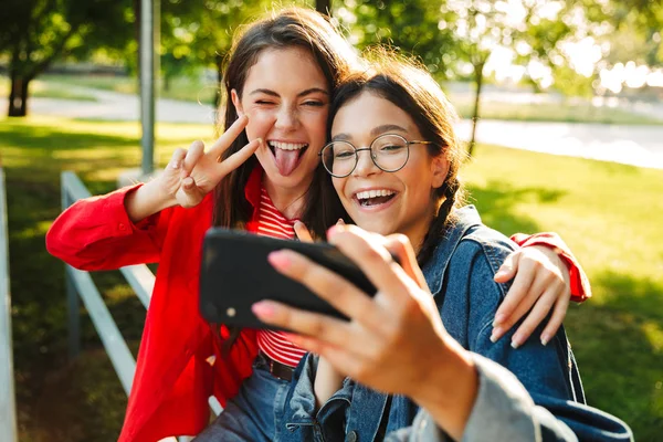 Imagen de dos chicas graciosas tomando fotos de selfies en el celular y haciendo gestos de paz cantando mientras están sentadas en la barandilla en el parque verde —  Fotos de Stock