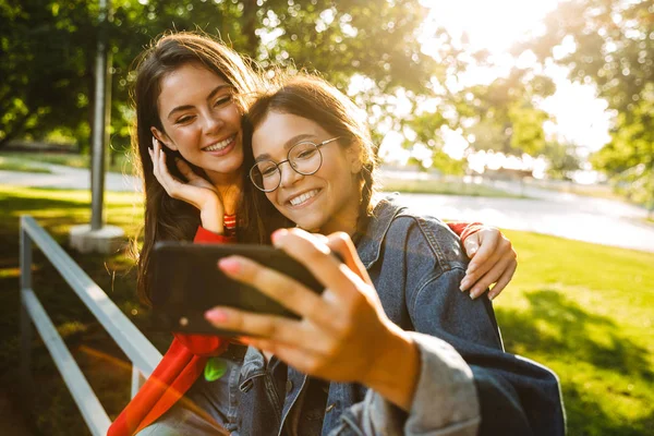 Yeşil parkta korkuluk üzerinde otururken cep telefonu selfie fotoğraf çekmek ve gülümseyen iki güzel kız görüntü — Stok fotoğraf