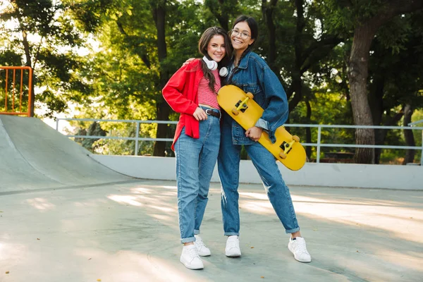 Imagen de dos chicas felices sonriendo y abrazándose juntas mientras sostienen el monopatín en el parque de skate —  Fotos de Stock