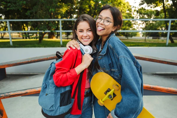 Imagem de duas meninas engraçadas sorrindo e abraçando juntos enquanto segurando skate no parque de skate — Fotografia de Stock