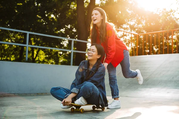 Imagem de duas meninas alegres rindo e andando de skate juntos no parque de skate — Fotografia de Stock