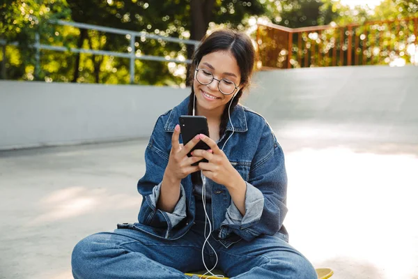 Kép csinos lány használ smartphone és fülhallgató ülve gördeszka a Skate Park — Stock Fotó