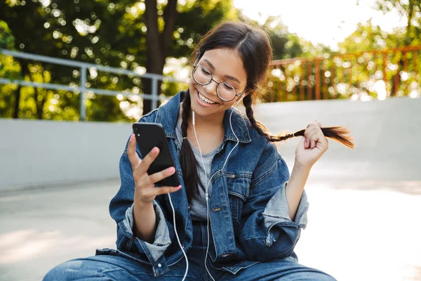 Kép a fiatal lány használ smartphone és fülhallgató ülve gördeszka a Skate Park — Stock Fotó