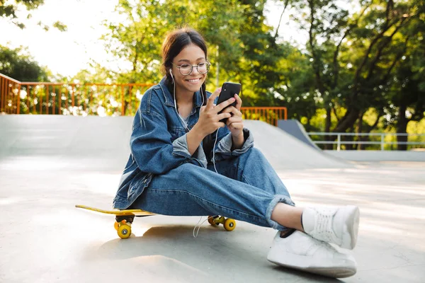 Bild av vacker flicka med hjälp av smartphone och hörlurar när du sitter på skateboard i Skate Park — Stockfoto