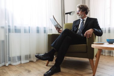 Olgun yakışıklı gri saçlı işadamı resmi kıyafetler içinde gazete okuma evde kapalı poz.