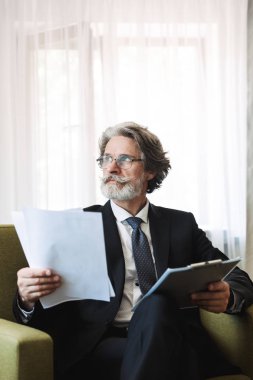 Serious handsome grey-haired senior businessman indoors at home dressed in formal clothes holding clipboard with paper documents.