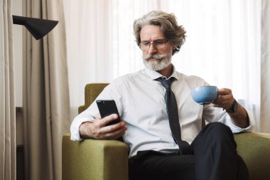 Konsantre ciddi düşünme üst düzey gri saçlı iş adamı evde kapalı sandalyede oturan akıllı telefon kahve içme kullanarak.