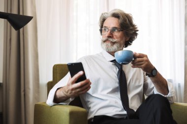 Konsantre ciddi düşünme üst düzey gri saçlı iş adamı evde kapalı sandalyede oturan akıllı telefon kahve içme kullanarak.