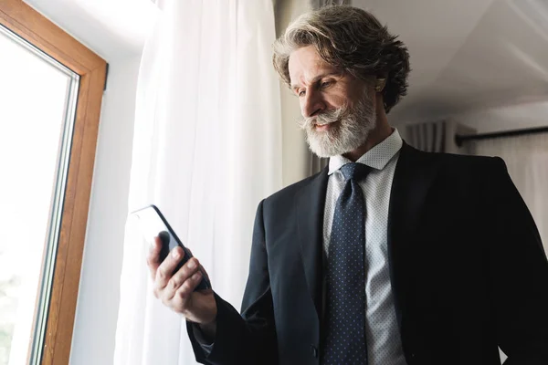 白人成年商人穿着黑色西装在酒店公寓中手持智能手机的照片 — 图库照片