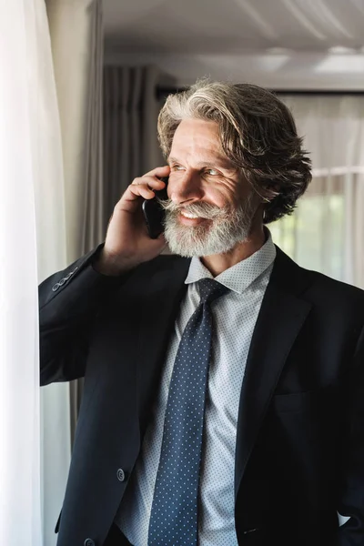 Image of happy adult businessman wearing black suit talking on smartphone in hotel apartment — Stock Photo, Image