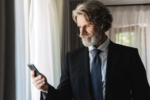 酒店公寓内使用智能手机和耳塞佩戴黑色西装的成人商人形象 — 图库照片