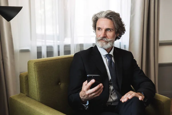 Красивый пожилой седой бизнесмен сидит в кресле дома, используя мобильный телефон. . — стоковое фото