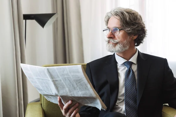 Mature handsome grey-haired businessman posing indoors at home dressed in formal clothes reading newspaper. — Stock Photo, Image
