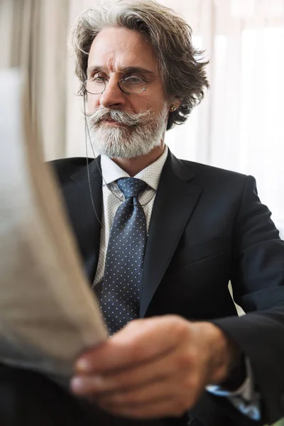 Concentrated handsome grey-haired senior businessman indoors at home dressed in formal clothes reading newspaper. — Stock Photo, Image