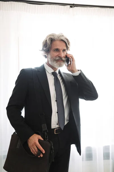 Zufriedene gutaussehende grauhaarige Senior-Geschäftsmann drinnen zu Hause in formeller Kleidung spricht mit Handy-Tasche. — Stockfoto