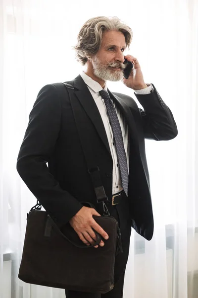 심각한 잘 생긴 회색 머리 고위 사업가 실내 에서 집에서 옷을 입고 공식적인 옷을 입고 말하는 휴대 전화 들고 가방. — 스톡 사진