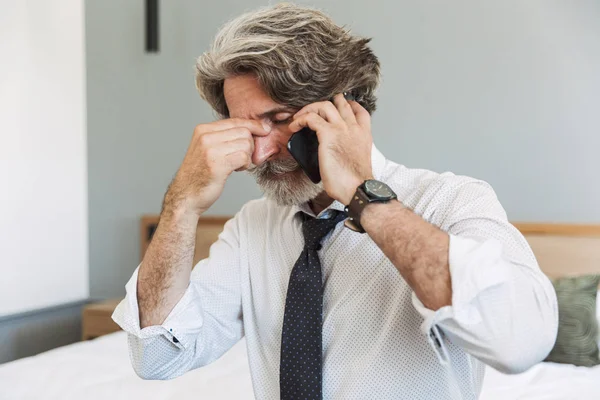Triste hombre de negocios de pelo gris mayor sentado en la cama en casa hablando por teléfono móvil . — Foto de Stock