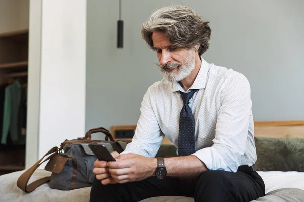 Seriózní starší šedovlasý obchodní muž, který sedí na posteli doma a používá mobilní telefon. — Stock fotografie