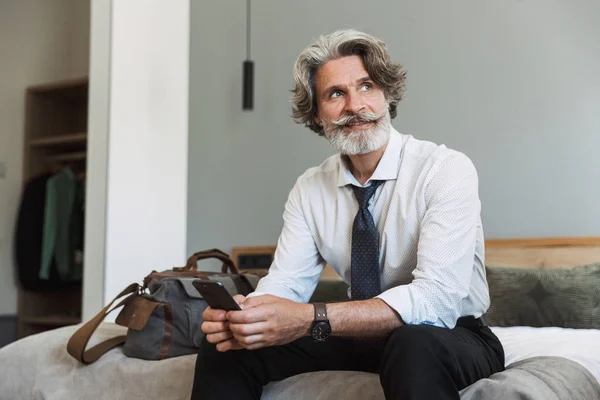 Älterer grauhaariger Geschäftsmann sitzt zu Hause mit Handy im Bett. — Stockfoto