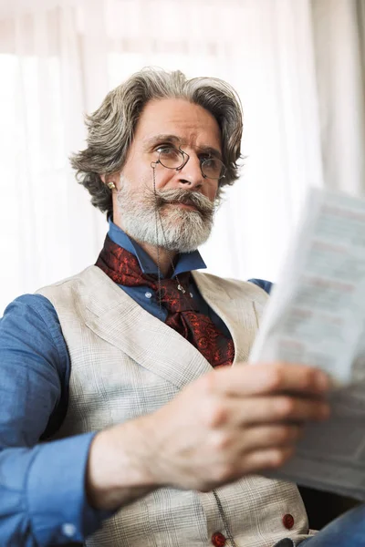 Portrait of serious adult businessman reading newspaper while sitting on armchair in hotel apartment — Stock Photo, Image
