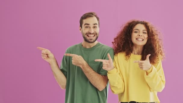 微笑积极的年轻夫妇指着一边用手指 使拇指向粉红色背景的手势隔离 — 图库视频影像