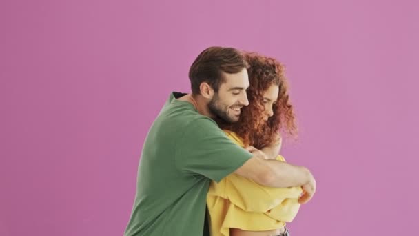 彼のガールフレンドを抱きしめる若いあごひげのブルネットの男を微笑む かわいい若い赤毛の巻き毛の女性は 彼女のボーイフレンドに動揺感じています ピンクの背景の上に美しい若いカップル孤立 — ストック動画