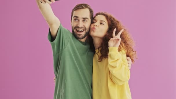 魅力的な幸せな若い素敵なカップルが抱きしめ ピンクの背景の上にスマートフォン上の自分撮り写真を作る孤立 — ストック動画