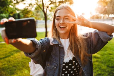 Doğa yeşil parkta yürüyen olumlu iyimser genç kız cep telefonu ile selfie çekmek.