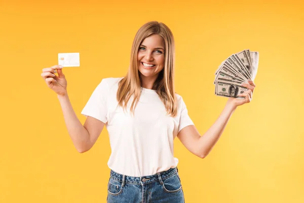 Imagen de primer plano de la mujer rubia positiva con camiseta casual regocijándose mientras sostiene la tarjeta de crédito y dinero en efectivo —  Fotos de Stock
