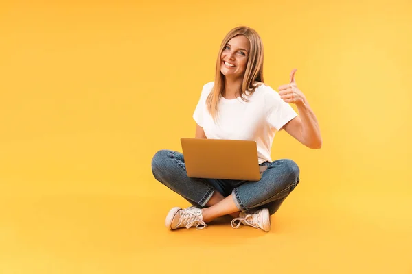 Imagem de mulher loira caucasiana mostrando polegar para cima e usando computador portátil enquanto sentado com as pernas cruzadas — Fotografia de Stock