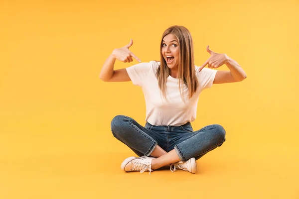 Imagem de mulher loira atraente sorrindo e apontando o dedo para si mesma enquanto sentada com as pernas cruzadas — Fotografia de Stock