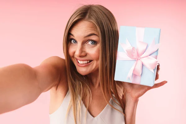 Image of charming young woman wearing dress smiling and holding gift box while taking selfie photo — Φωτογραφία Αρχείου