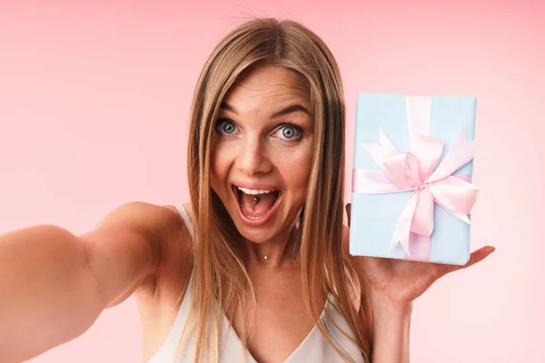 Εικόνα της ενθουσιασμένες νεαρή γυναίκα φορώντας φόρεμα εκφράζοντας έκπληξη και κρατώντας κουτί δώρου ενώ παίρνετε selfie φωτογραφία — Φωτογραφία Αρχείου