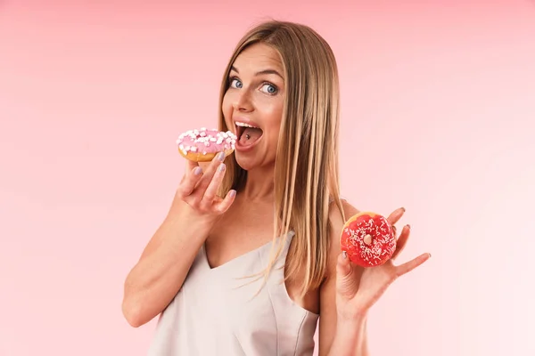 Immagine di attraente donna bionda che indossa un vestito sorridente mentre mangia due ciambelle dolci — Foto Stock