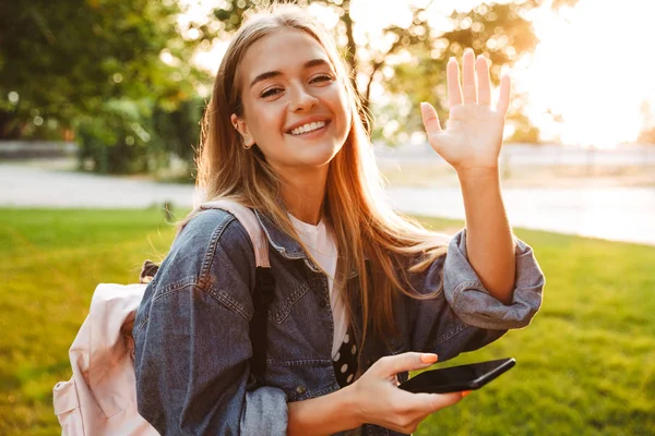 Meisje wandelen buiten in natuur groen park zwaaiend naar u met behulp van mobiele telefoon. — Stockfoto