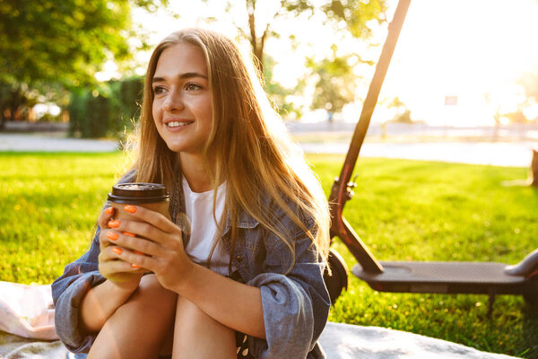 Cheery smiling young teenage girl on grass with scooter drinking coffee.