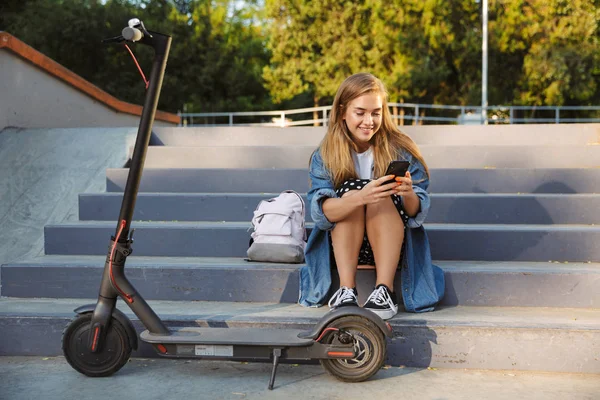 Positiv glada tonårs flicka i Park sitta på stammen nära Scooter användande rörlig telefonerna. — Stockfoto