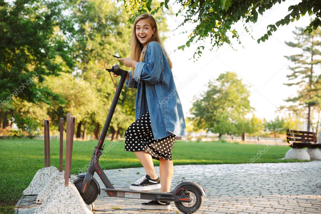Happy teenage girl in park walking on scooter looking camera.