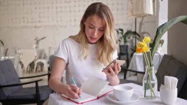 Akıllı telefon kullanan ve kafede otururken kalemle not defterine yazan konsantre olmuş sarışın bir kadın.