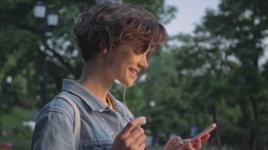 Kot ceketli, gülümseyen, kumral bir kadın, kulağına kulaklık takıyor ve parkta akıllı telefon kullanırken el sallıyor.