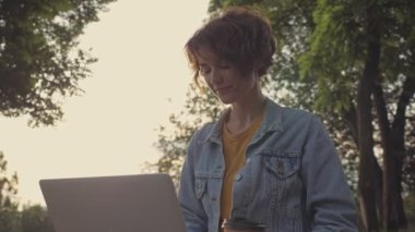 Parkta dizüstü bilgisayarla çalışırken kot ceketli, gülümseyen güzel, esmer bir kadın.