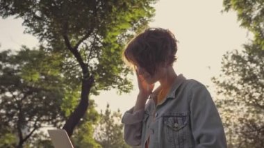 Kot ceketli, güzel, esmer bir kadının yan görünüşü. Parkta dizüstü bilgisayarlı bir kulaklıkla gülüyor ve el sallıyor.