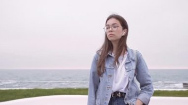 Kot ceketli esmer kız deniz kenarında kalırken etrafa bakıyor ve derin derin nefes alıyor.