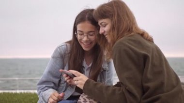 Neşeli genç kız arkadaşlar akıllı telefondan bir şeyler izleyip deniz kenarındaki iskelede otururken gülüyorlar.