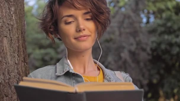Привлекательная Юная Брюнетка Джинсовой Куртке Слушает Музыку Наушниками Читает Книгу — стоковое видео
