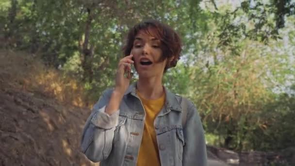 魅力的な陽気な若いですブルネット女性でデニムジャケットスマートフォンで話している間に公園を歩く — ストック動画