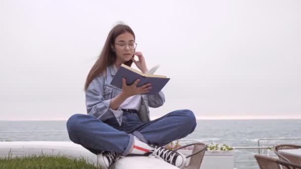 坐在靠近大海的码头边看书时 全神贯注地穿着牛仔裤和牛仔夹克的年轻黑发姑娘 — 图库视频影像