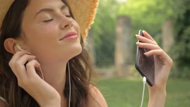 在公园里 你可以近距离地看到戴着草帽的快乐的年轻黑发女人坐在公园里 一边笑一边用智能手机听音乐 — 图库视频影像