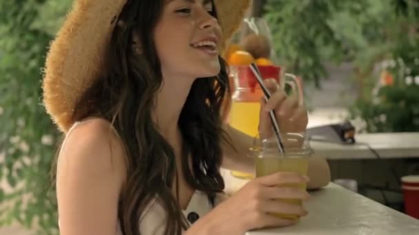 かわいいです陽気な若いですブルネット女性でわら帽子飲む新鮮なジュースからプラスチックカップしばらく座っている間バーカウンターでザ公園 — ストック動画
